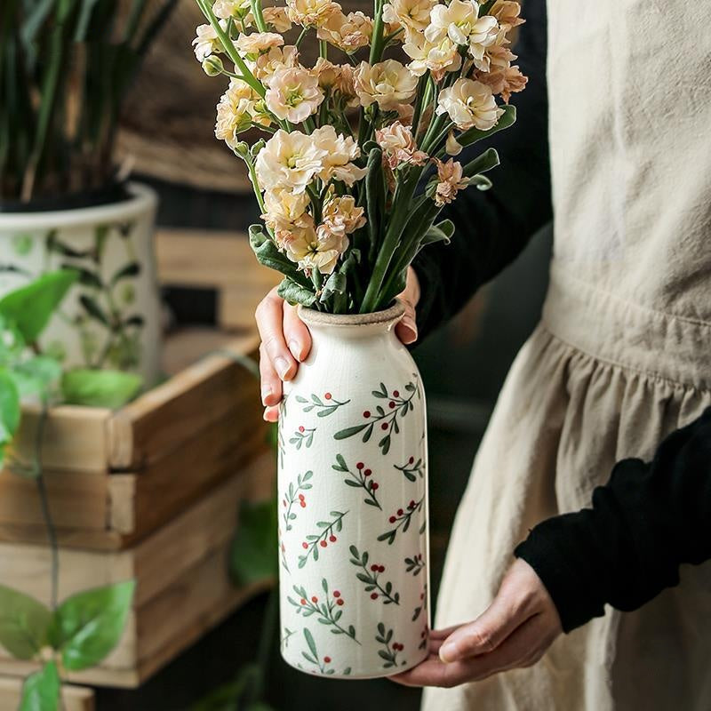 Person holding a white vase with floral patterns filled with flowers, blurred indoor background
