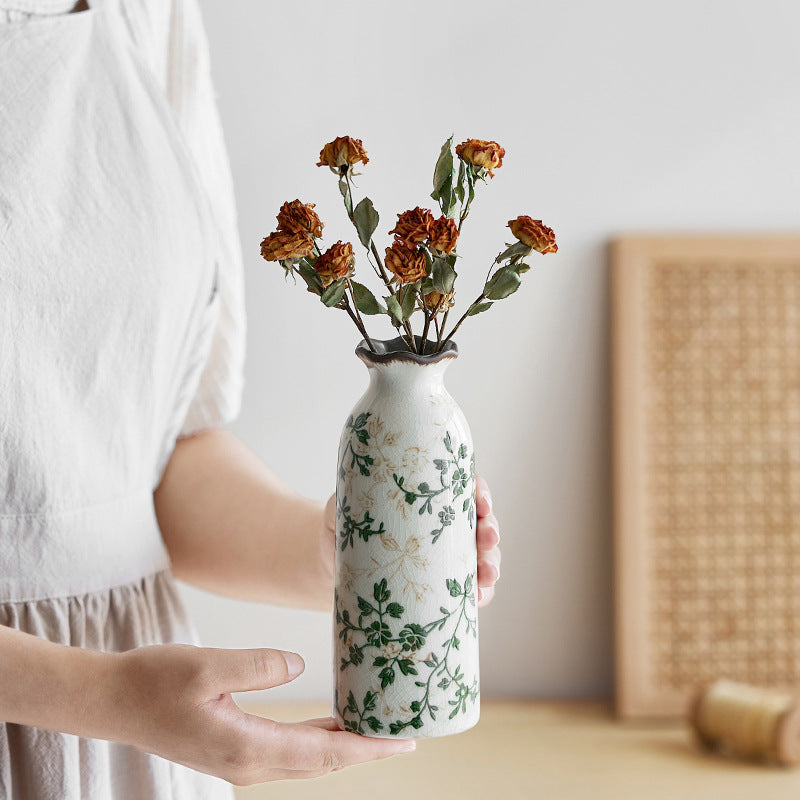 Person holding a white vase with floral design and dried flowers against a neutral background