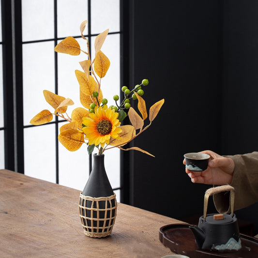 Black vase with sunflower and leaves on a wooden table, hand holding a cup in the background