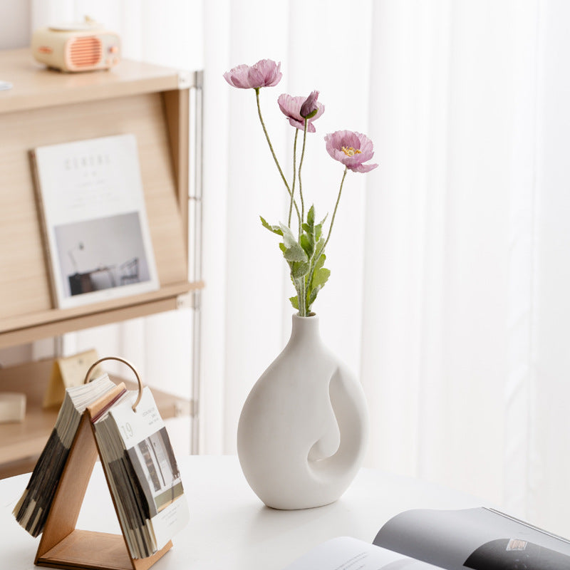 White vase with pink flowers on a table next to a bookshelf