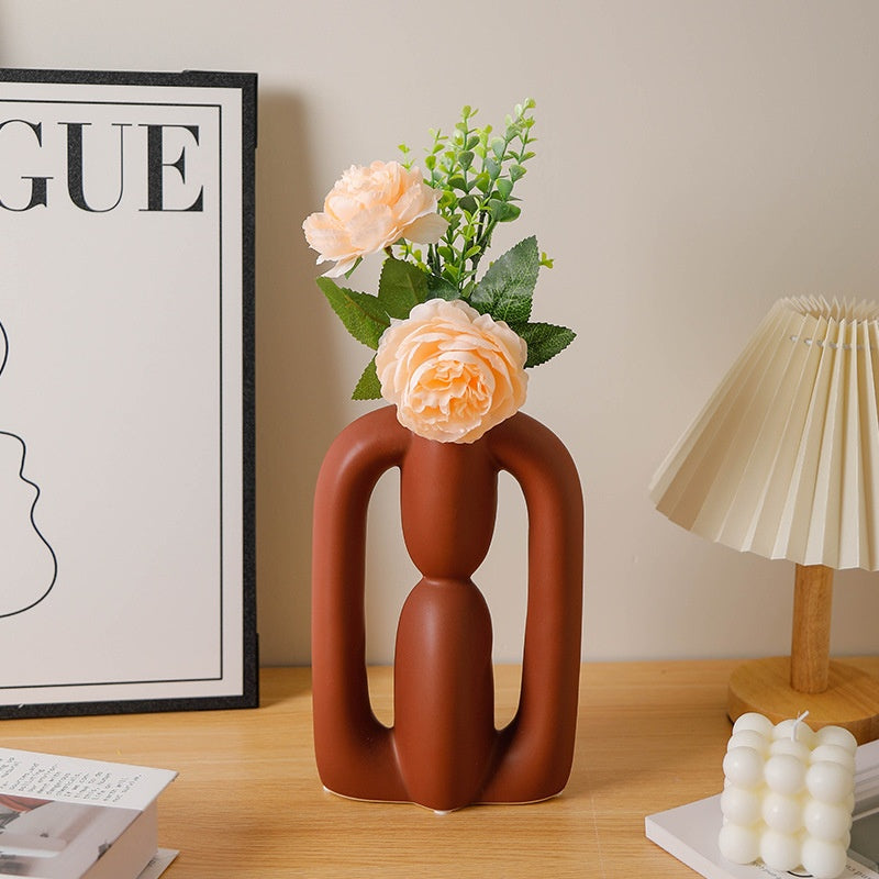 Abstract brown vase with flowers on a wooden surface next to a lamp and framed poster.