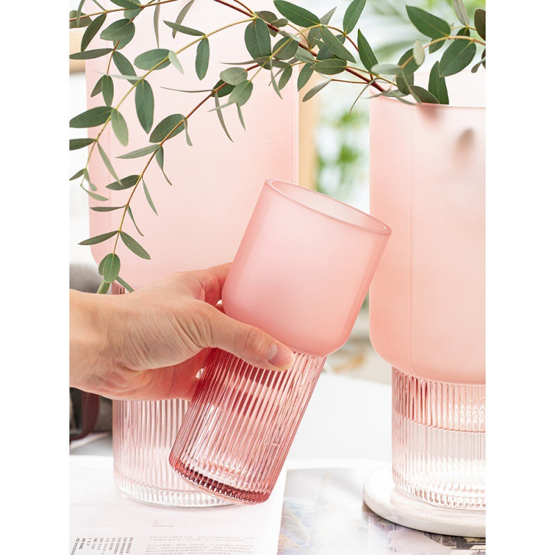 Pink glass tumblers with ribbed design held by a hand, against a blurred background with greenery.