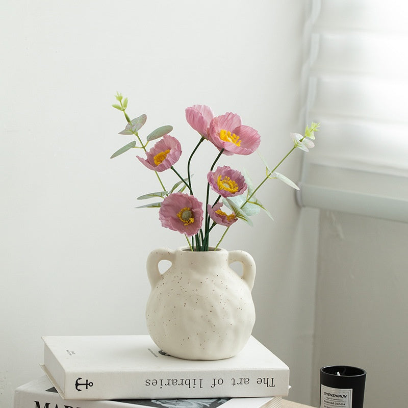 White ceramic vase with pink flowers on a stack of books against a white wall.