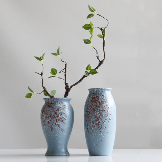 Two blue ceramic vases with speckled patterns holding branches with green leaves on a light gray background.