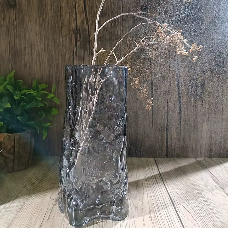 Decorative glass vase with textured surface on a wooden floor, with plants in the background.