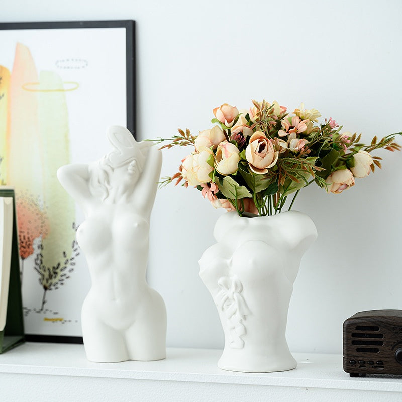 White ceramic vase shaped like a face with a small figurine on top, filled with flowers on a white surface.