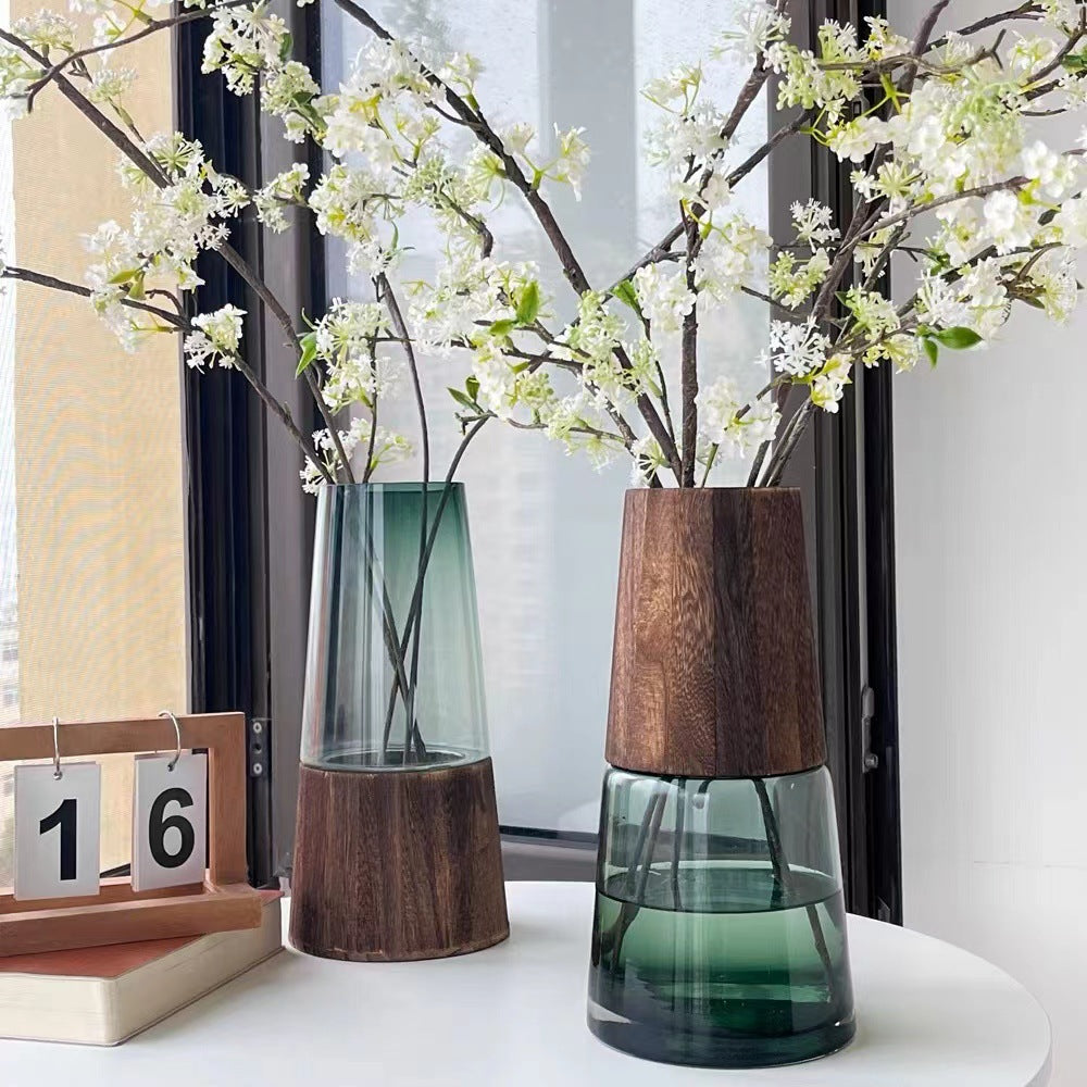Two vases with wooden bases and glass bodies, one containing white flowers, on a white surface.