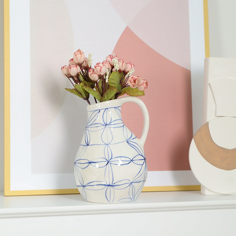 White vase with blue patterns holding pink flowers on a shelf with a abstract art piece in the background