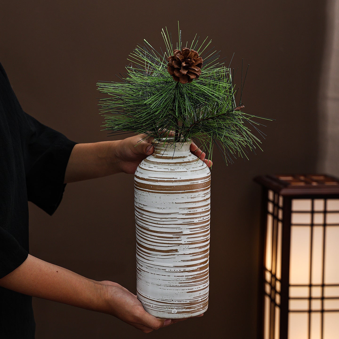 Person holding a textured white vase with greenery and a pine cone against a dark background