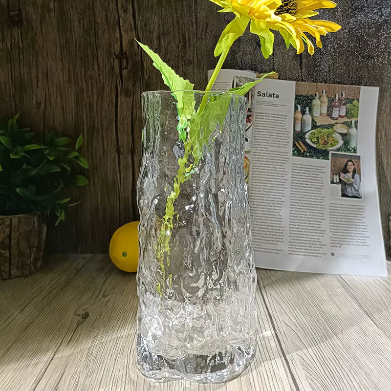 Clear textured vase with a yellow flower on a wooden surface next to a magazine.