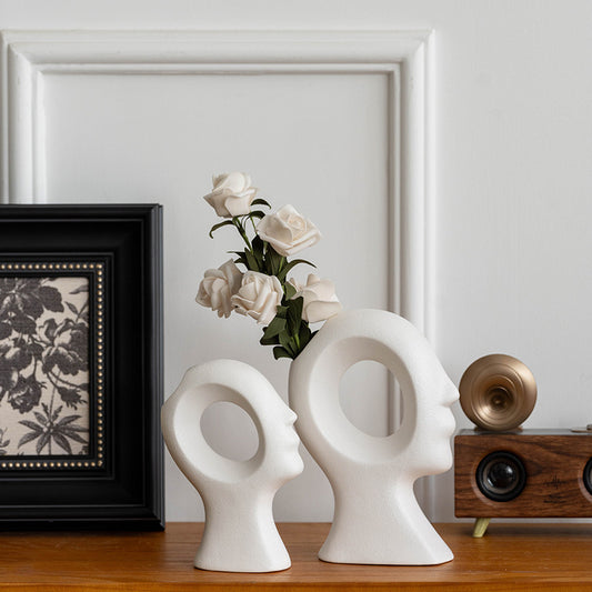 Two white face-shaped vases on a wooden surface with a framed picture and decorative items in the background.