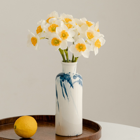 White vase with blue abstract design containing white flowers with yellow centers on a wooden tray.