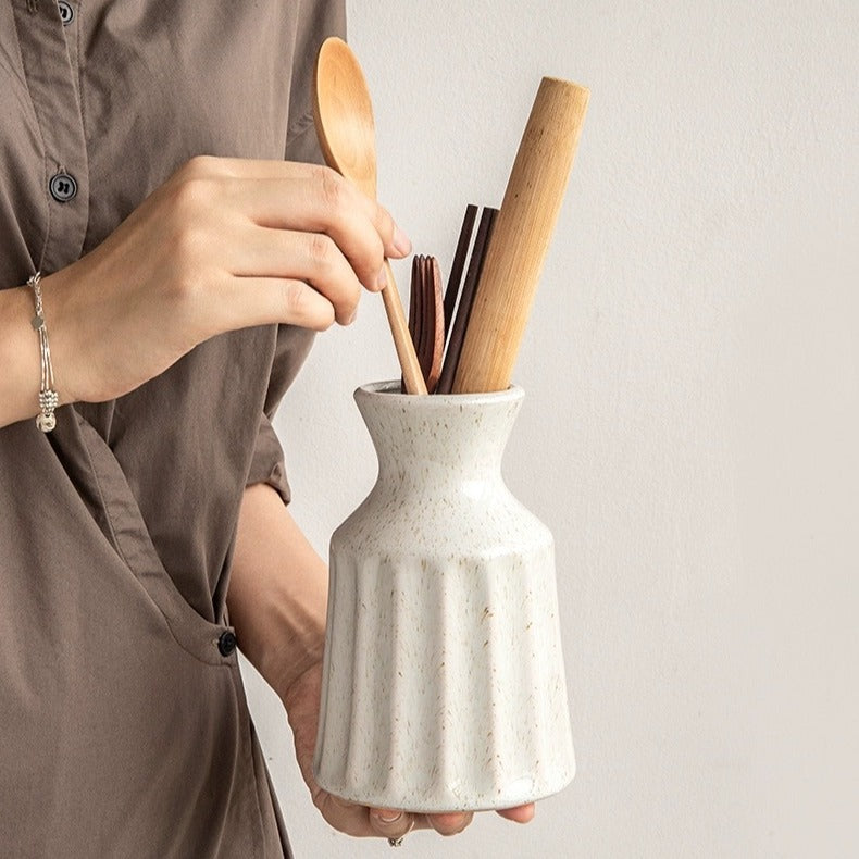 Person holding a white ceramic utensil holder with wooden spoons and chopsticks.