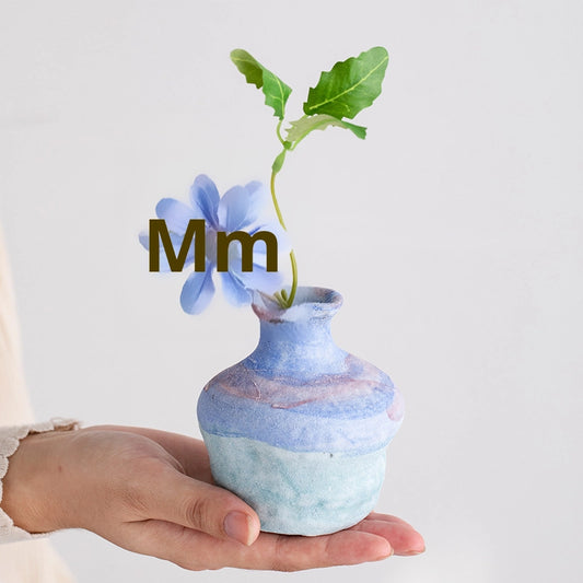 Hand holding a small blue and green ceramic vase with a flower, on a light gray background.