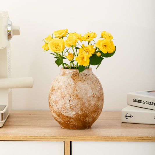 Decorative vase with yellow flowers on a wooden shelf against a white wall.