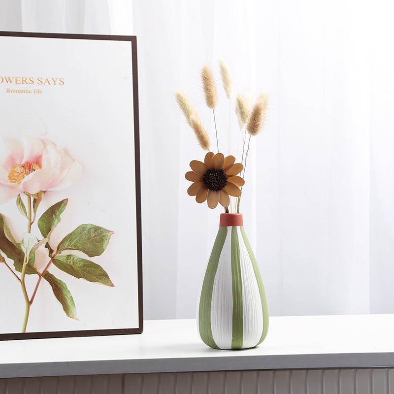 Green and white vase with dried flowers on a surface, with a floral-themed painting in the background.