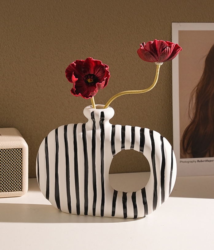 Black and white striped vase with red flowers on a shelf against a brown wall.