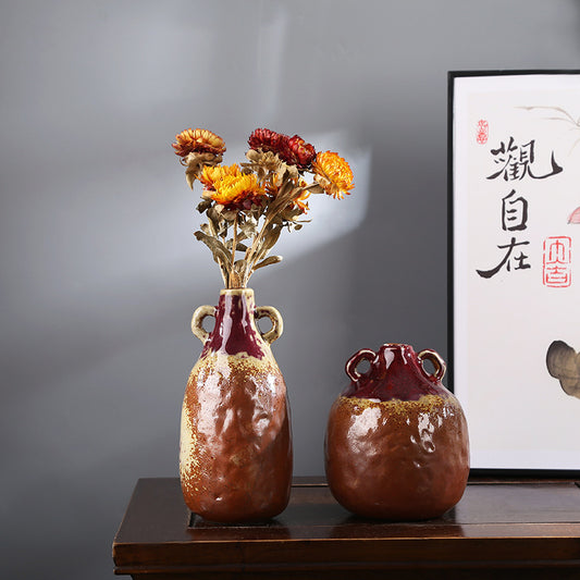 Two ceramic vases with dried flowers on a wooden surface against a gray wall with a framed picture.