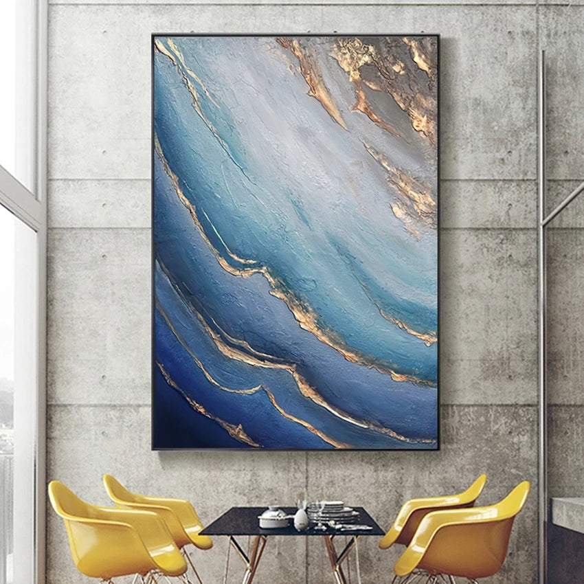 Abstract painting with blue and gold tones on a concrete wall above a dining table with yellow chairs.