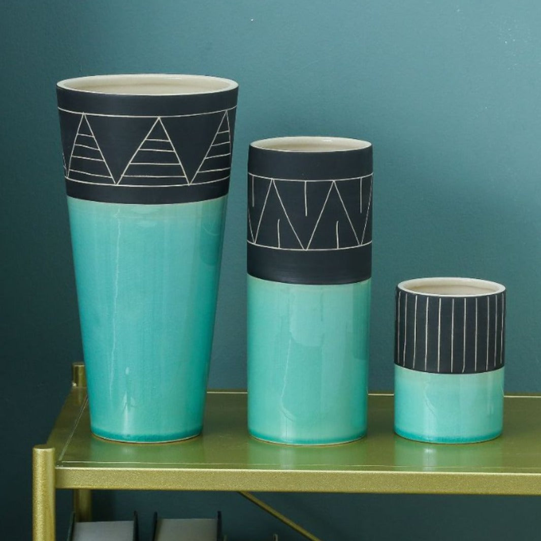 Three turquoise vases with black geometric designs on a green surface against a teal wall.