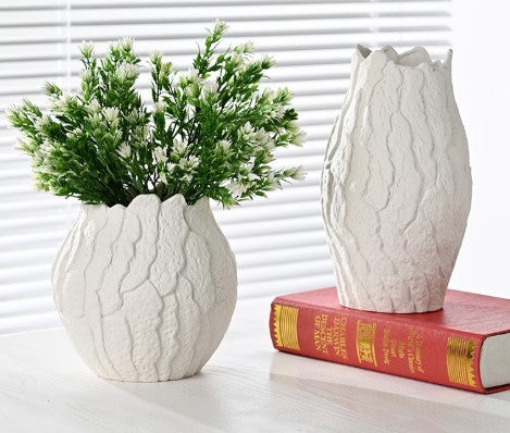 Two white textured vases with one holding greenery on a white surface.