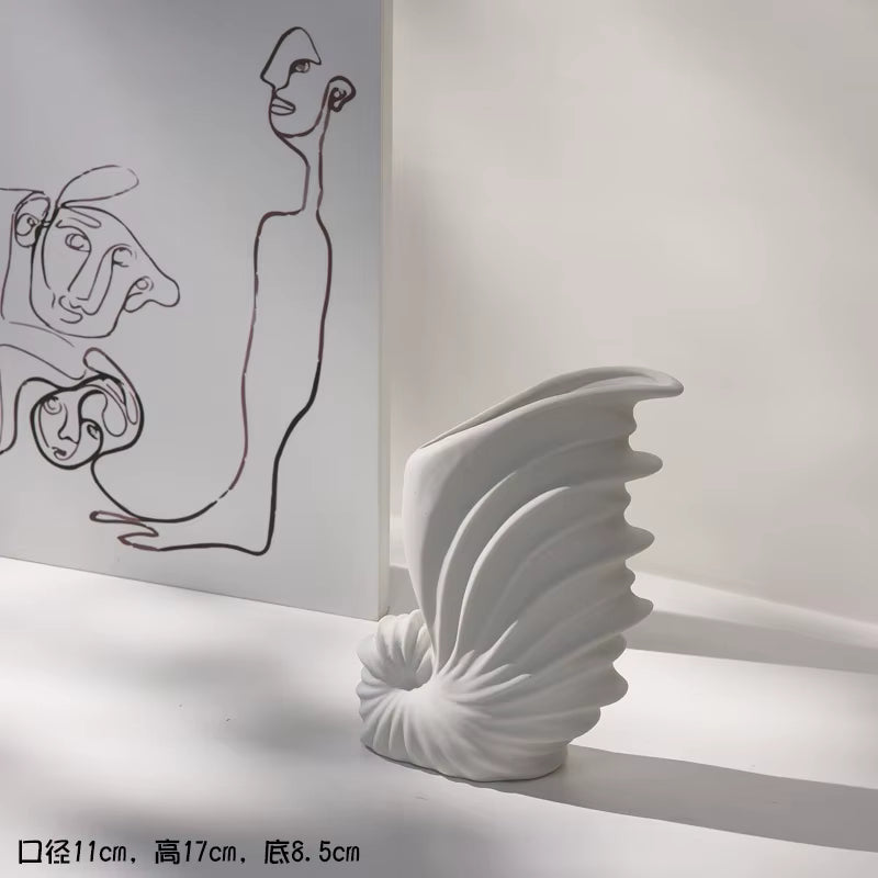 White ceramic vase with shell design on a white surface, with abstract line art in the background.