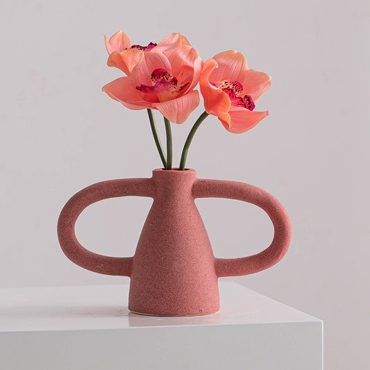 Pink vase with handles holding pink flowers on a white surface with a light gray background