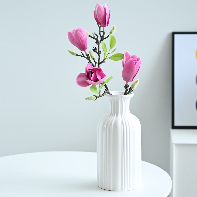 White vase with pink flowers on a white surface against a light gray wall.