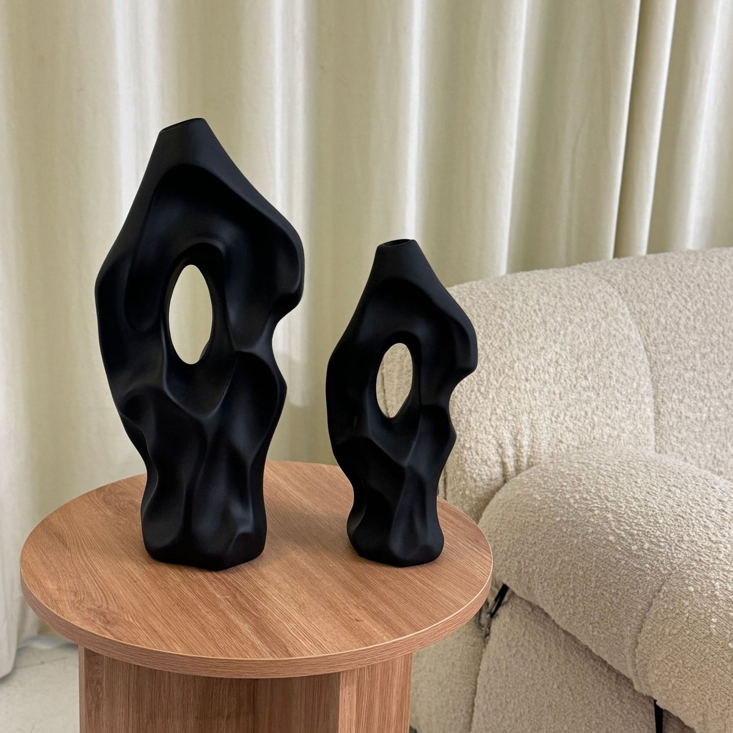 Two black abstract sculptures on a wooden table with a beige sofa and curtain in the background.