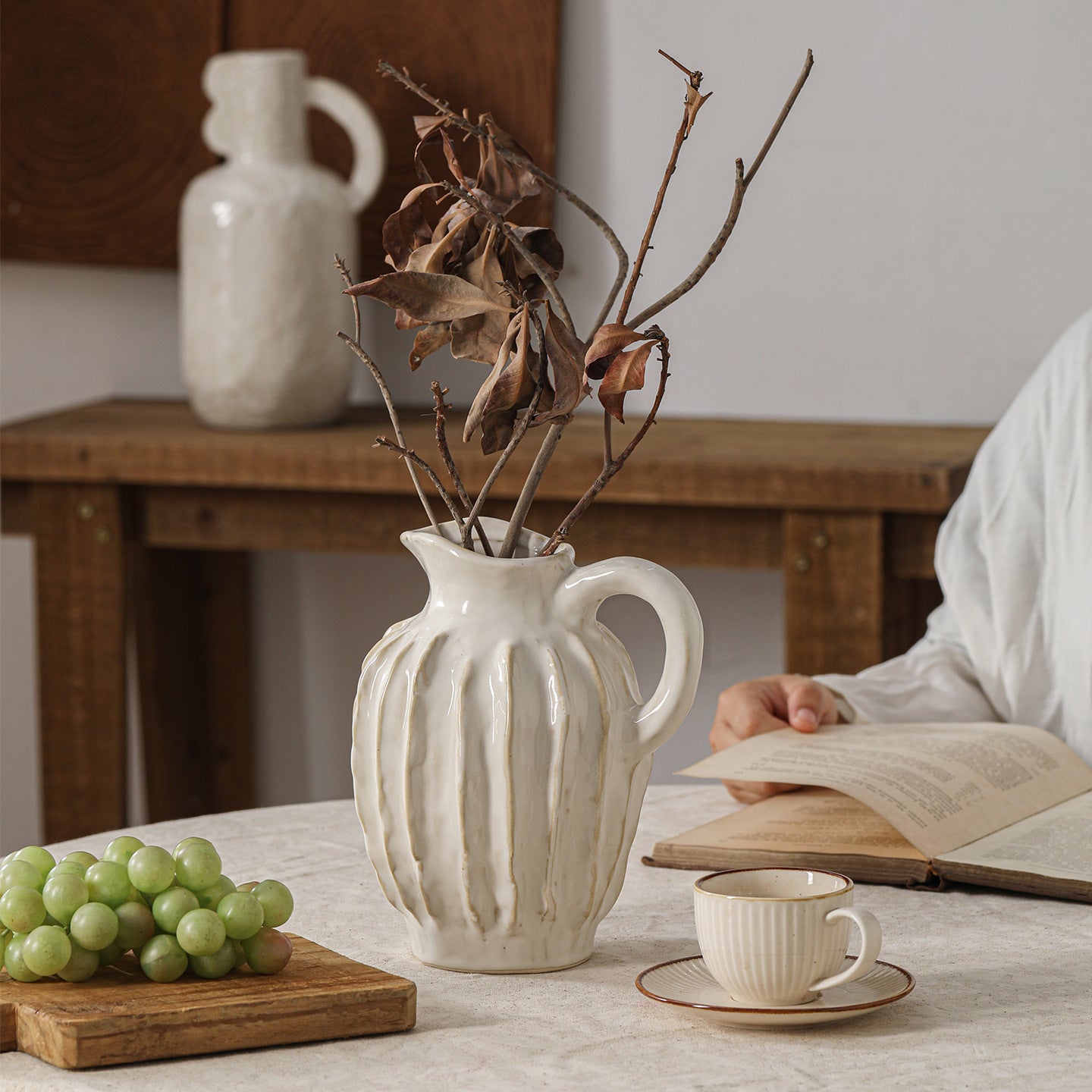 White ceramic pitcher with dried branches on a table with grapes and a book