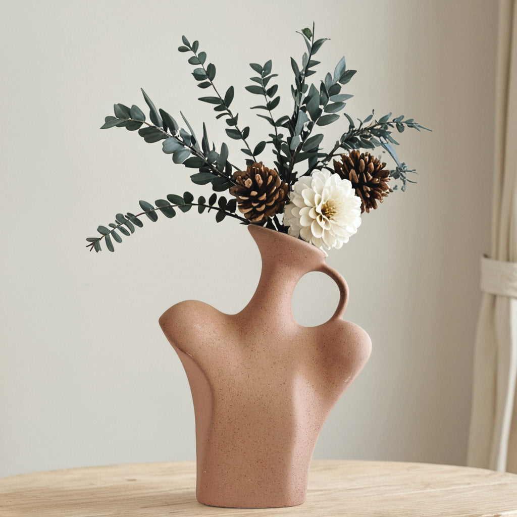 Ceramic vase with a unique design filled with greenery, pinecones, and a white flower on a wooden surface.