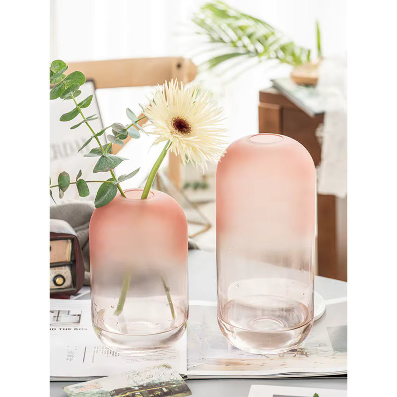 Two gradient pink vases with a single white flower on a table with a magazine and plant in the background.