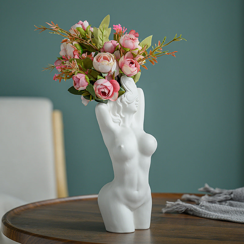 White ceramic vase shaped like a female torso with pink flowers on a wooden table.