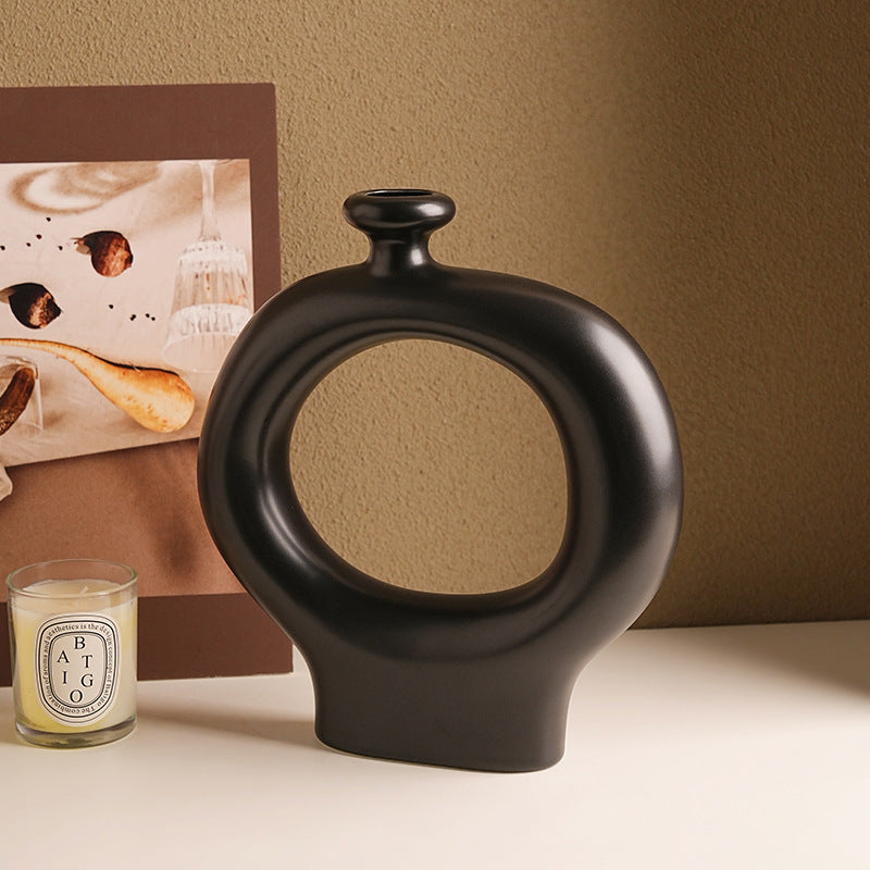 Black decorative vase with a circular design on a beige surface next to a framed picture and a candle.