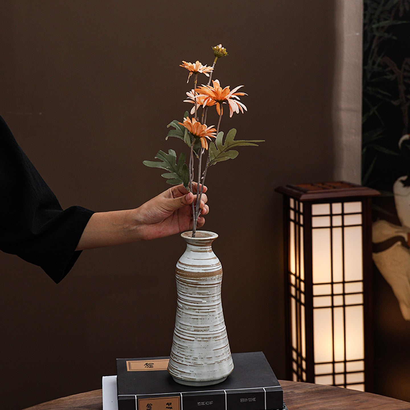 Person arranging flowers in a textured vase with a warm light lamp in the background