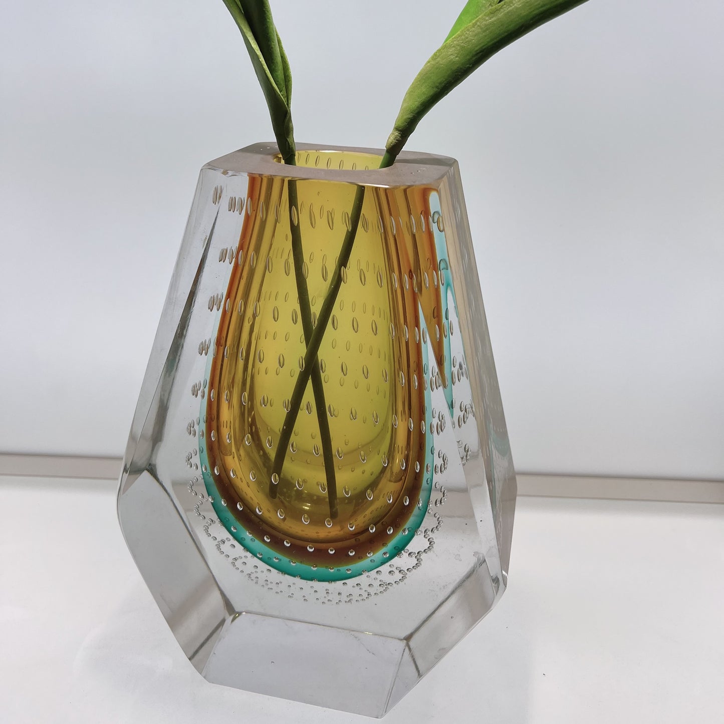 Geometric glass vase with a textured yellow center and clear sides, containing green leaves.
