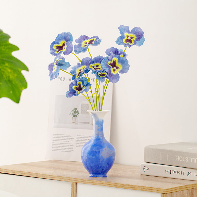 Blue and yellow flowers in a vase on a wooden table with books and a plant in the background.