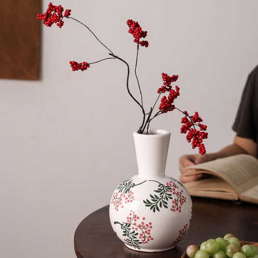 White vase with floral design and red berries on a table, with a person reading a book in the background.