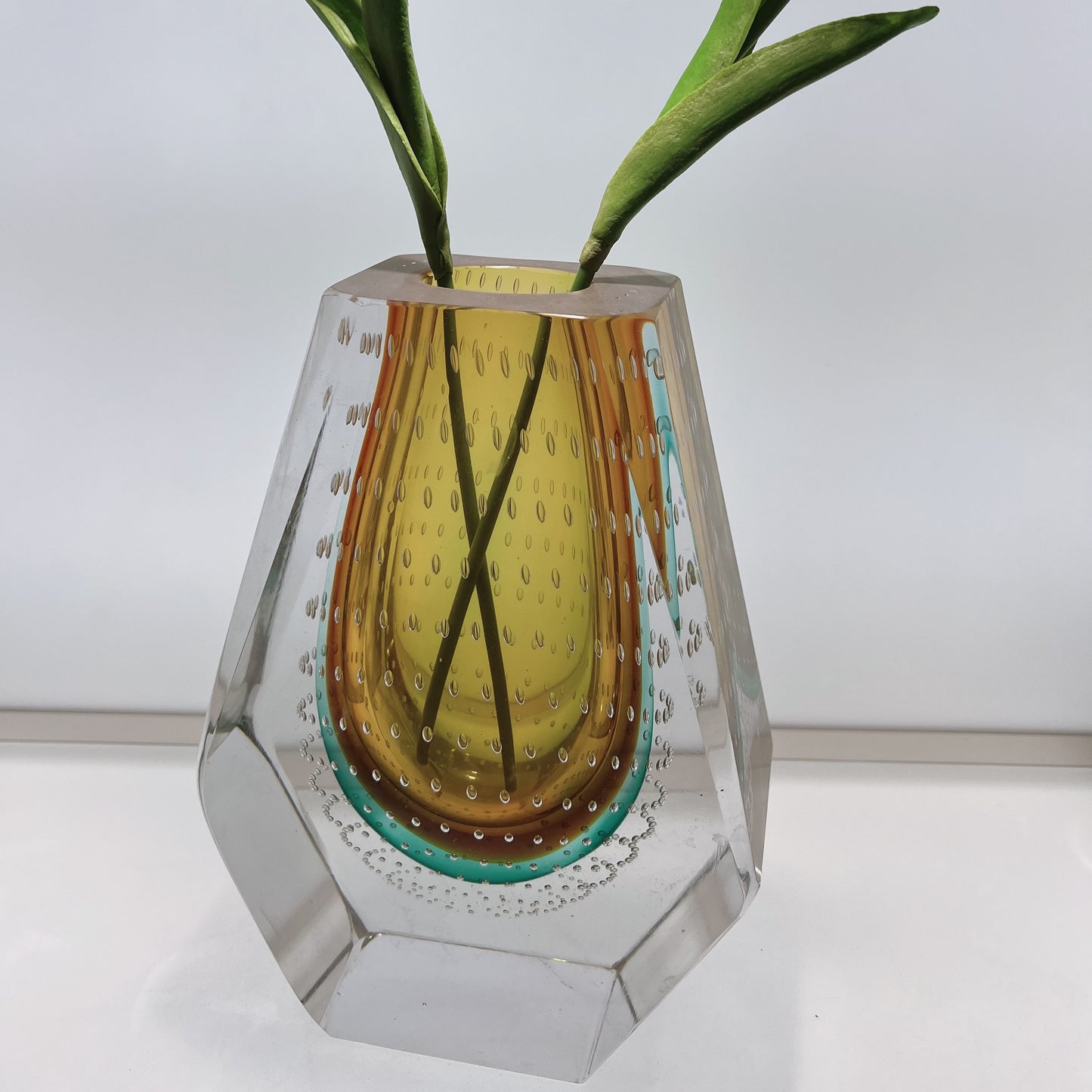 Glass vase with a yellow interior and clear exterior, containing green leaves on a white background