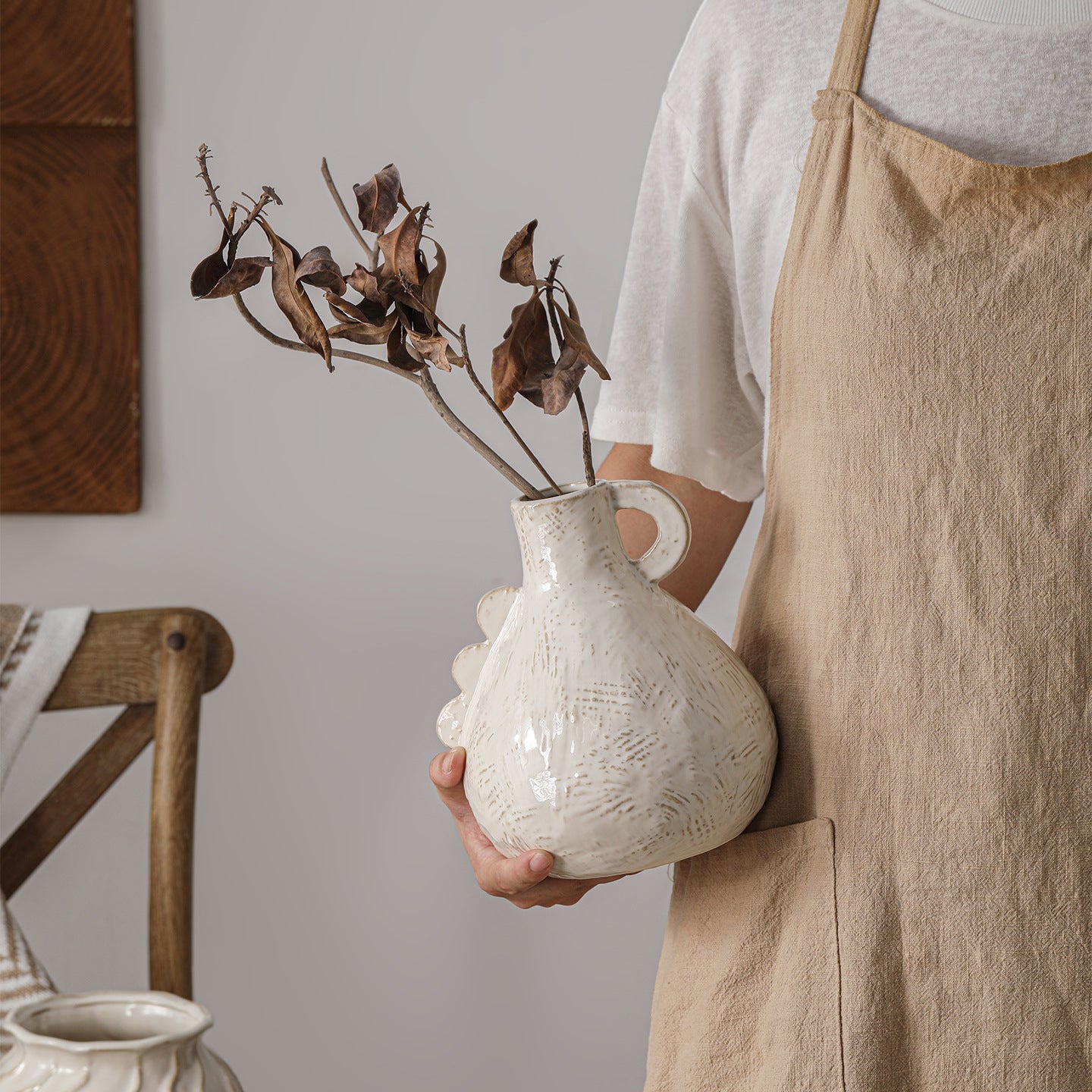 Person holding a textured white vase with dried branches indoors