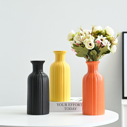 Three vases of different colors (black, yellow, orange) with a bouquet of flowers on a white surface.