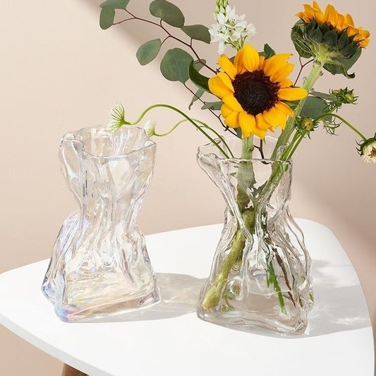 Two clear glass vases on a white surface with one containing sunflowers and greenery.