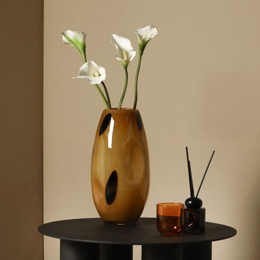 A hand-blown amber glass vase, inserted with four white lilies, sits on a black round table next to a glass mug and a defuser.