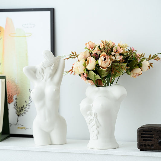 White ceramic vase shaped like a face with a small figurine on top, filled with flowers on a white surface.