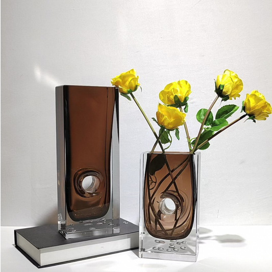 Two glass vases with a brown and clear design, one containing yellow flowers on a white background.