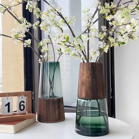 Two vases with wooden bases and glass bodies, one containing white flowers, on a white surface.