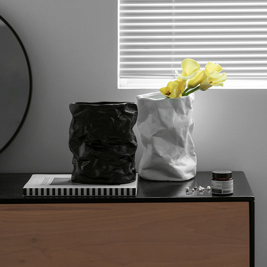 Two vases, one black and one white with yellow flowers, on a dark surface with a gray wall and window blinds in the background.