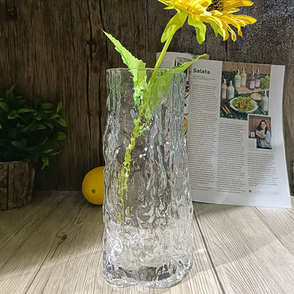 Clear textured vase with a yellow flower on a wooden surface next to a magazine.