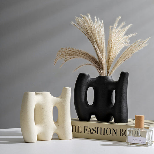 Two abstract vases, one black and one beige, with dried plants on a gray surface.