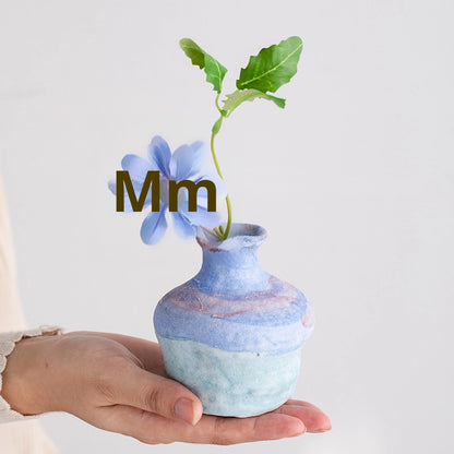 Hand holding a small blue and green ceramic vase with a flower, on a light gray background.